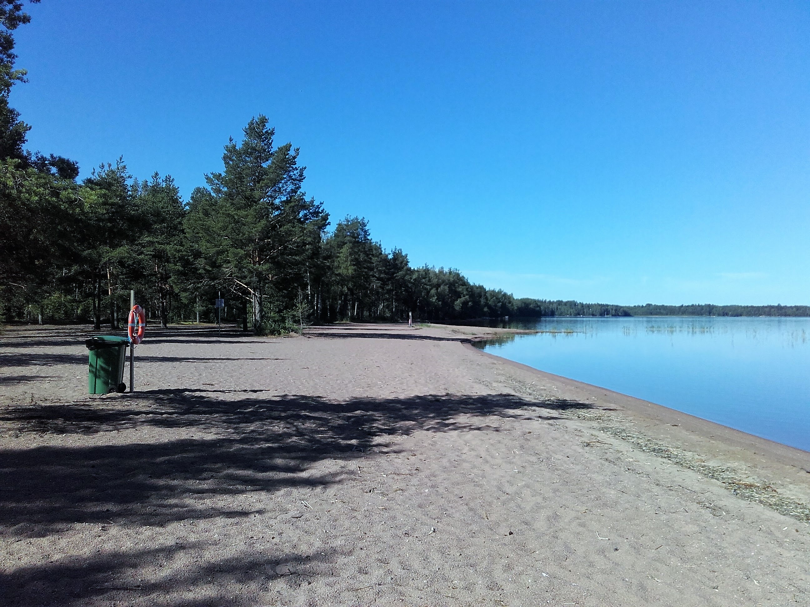 Särkkien uimaranta. Tasainen hiekkaranta. Hiekkarannalla vihreä roskakori ja telineessä pelastusrengas